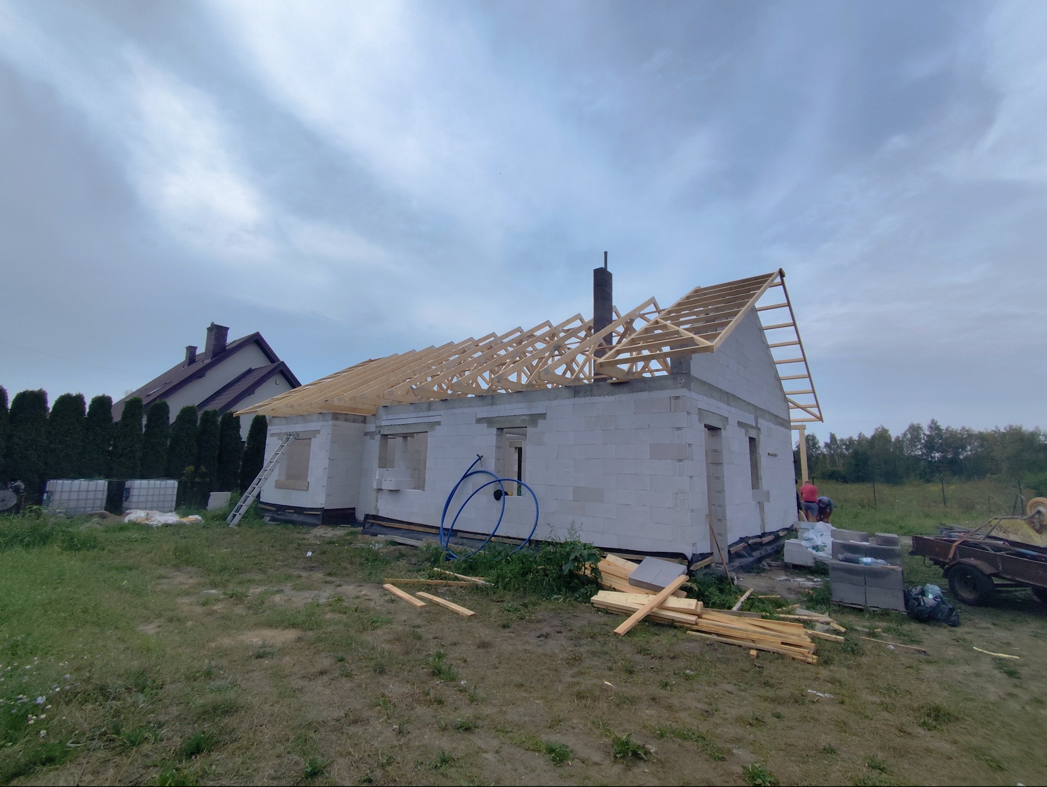 Budowa domu jednorodzinnego w Sadowne na etapie murowania ścian i konstrukcji dachu, widoczne białe bloczki, drewniana więźba dachowa, oraz materiały budowlane na posesji.