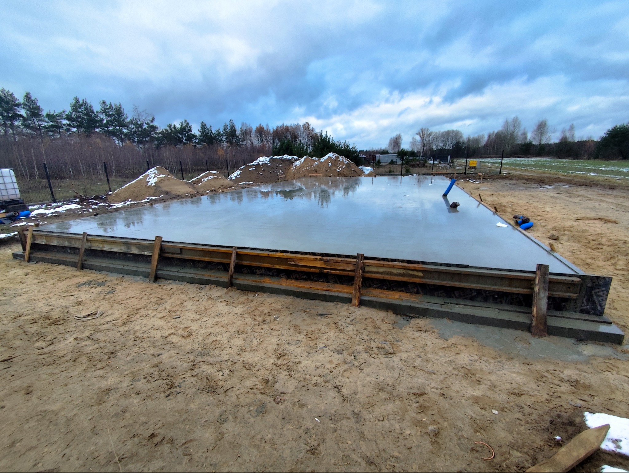 Świeżo wylany fundament z drewnianym szalunkiem, widoczne hałdy piasku z resztkami śniegu i pochmurne niebo w tle.