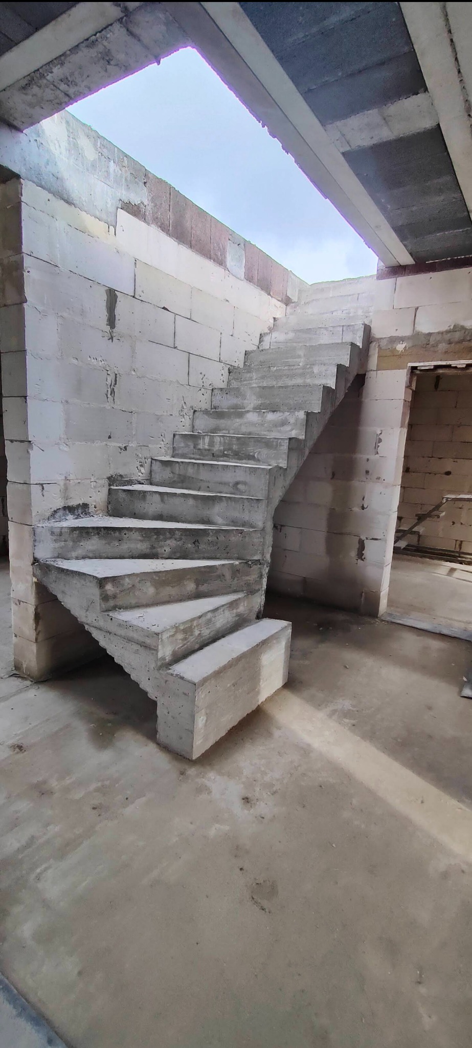 Surowe, betonowe schody spiralne w trakcie budowy, widoczne ściany z bloczków betonowych i surowy betonowy strop, brak wykończenia.