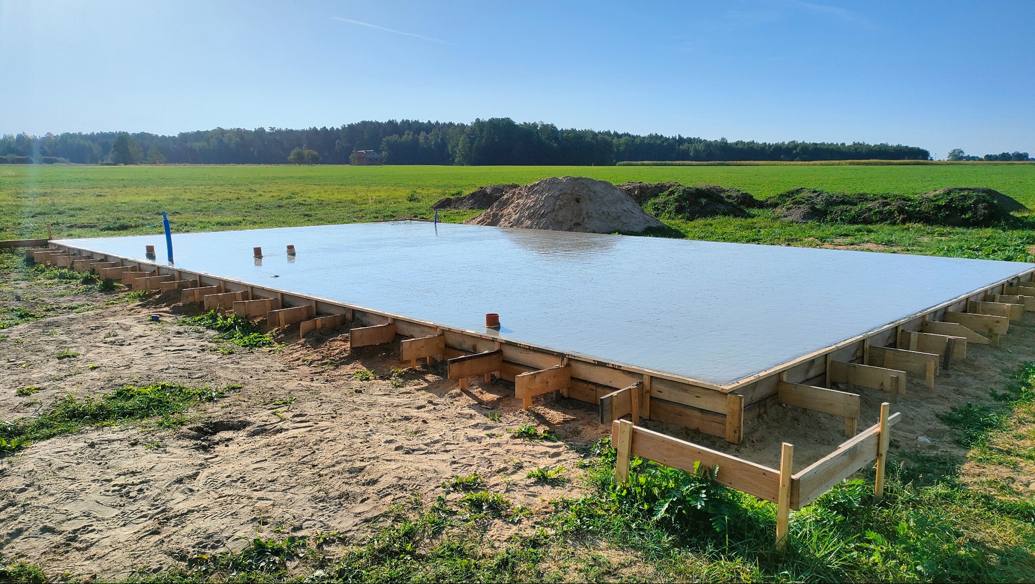 Świeżo wylany, gładki fundament betonowy z drewnianym szalunkiem, wystającymi rurami instalacyjnymi, na tle zielonego pola i lasu.