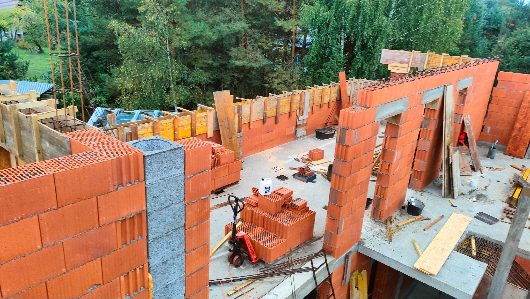 Wysoki kąt widzenia na budowę domu z czerwonej cegły Porotherm, z widocznymi elementami szalunków, zbrojenia i paletą z cegłami oraz wózkiem paletowym, otoczona drzewami.