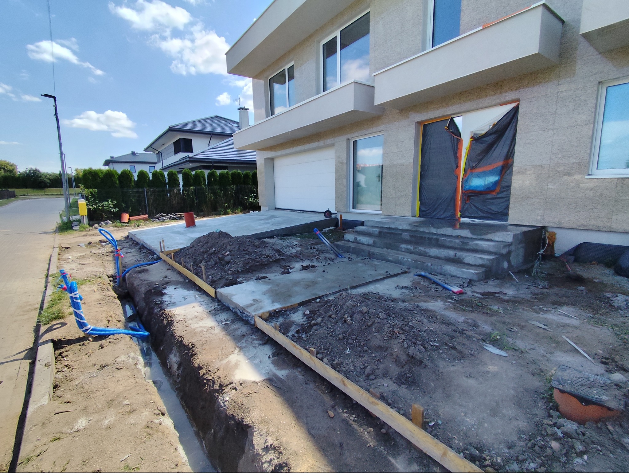 Nowoczesny dom w trakcie budowy, widoczne fundamenty z szalunkiem, podłączenia instalacji wodnej, zasłonięte wejście.