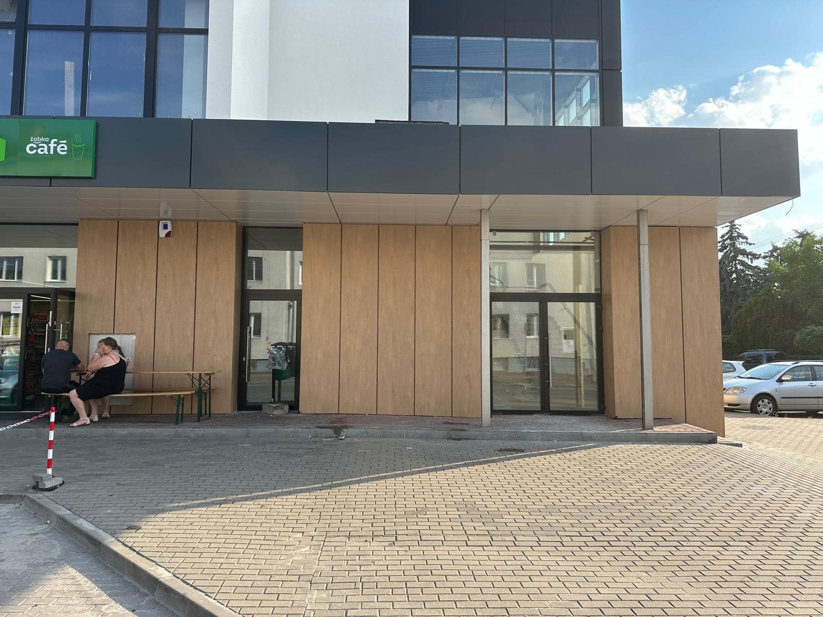 Fasada budynku z panelami imitującymi drewno, widoczne szklane drzwi wejściowe i okna, na zewnątrz siedzą ludzie na ławce.