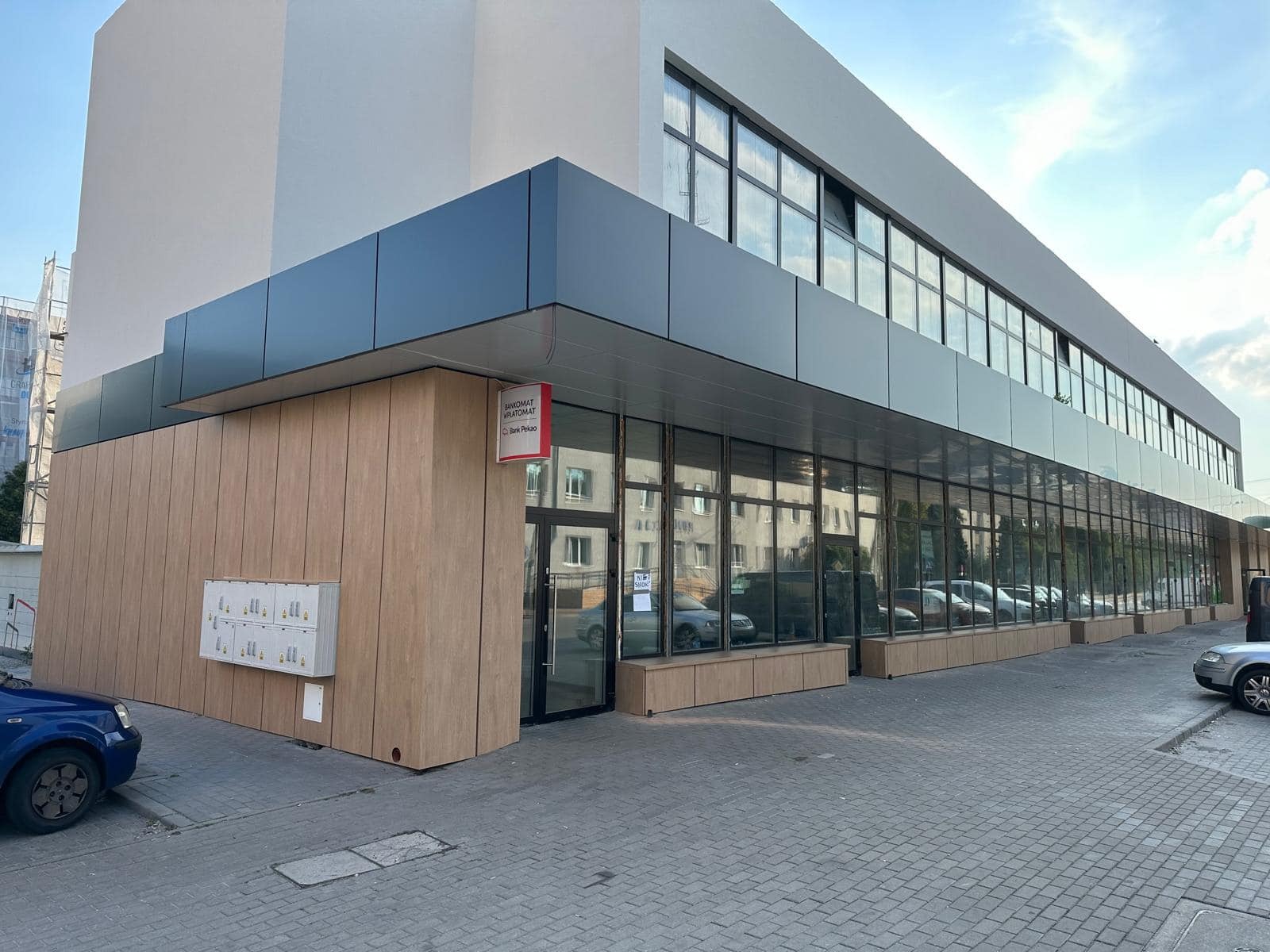 Nowoczesny budynek biurowy z elewacją wykończoną panelami w odcieniach szarości i drewna, duże okna, skrzynki pocztowe i szyld bankomatu przy wejściu.
