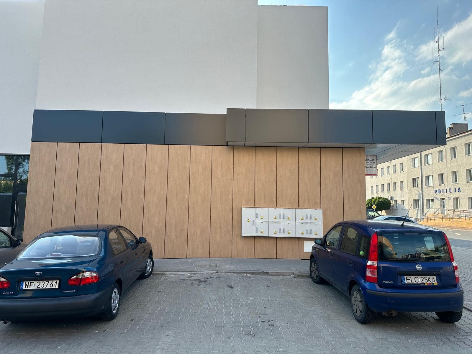 Nowoczesna elewacja budynku z drewnianymi panelami i szarym zadaszeniem, widoczne skrzynki pocztowe, zaparkowane samochody i budynek policji w tle.