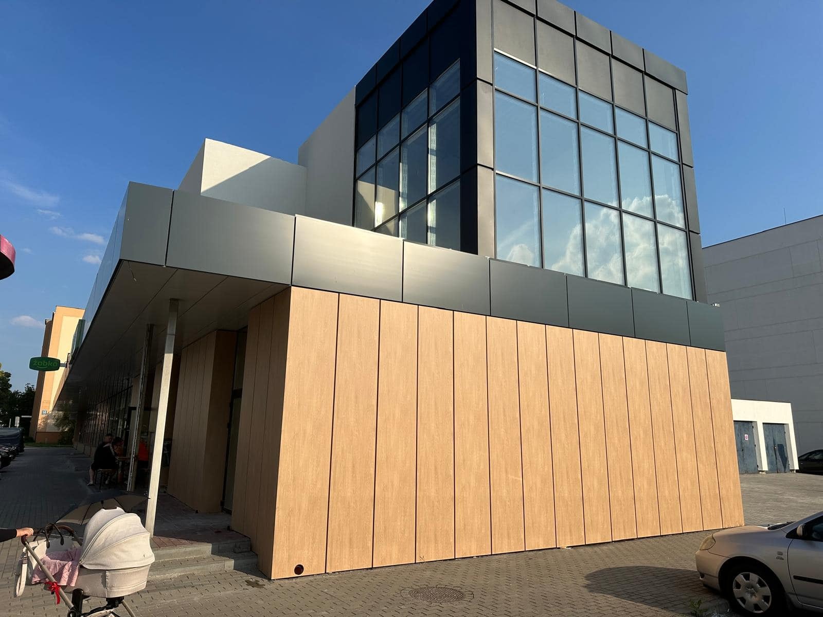 Nowoczesny budynek handlowy z elewacją z paneli drewnopodobnych i dużymi oknami, widok z poziomu chodnika, w tle błękitne niebo.