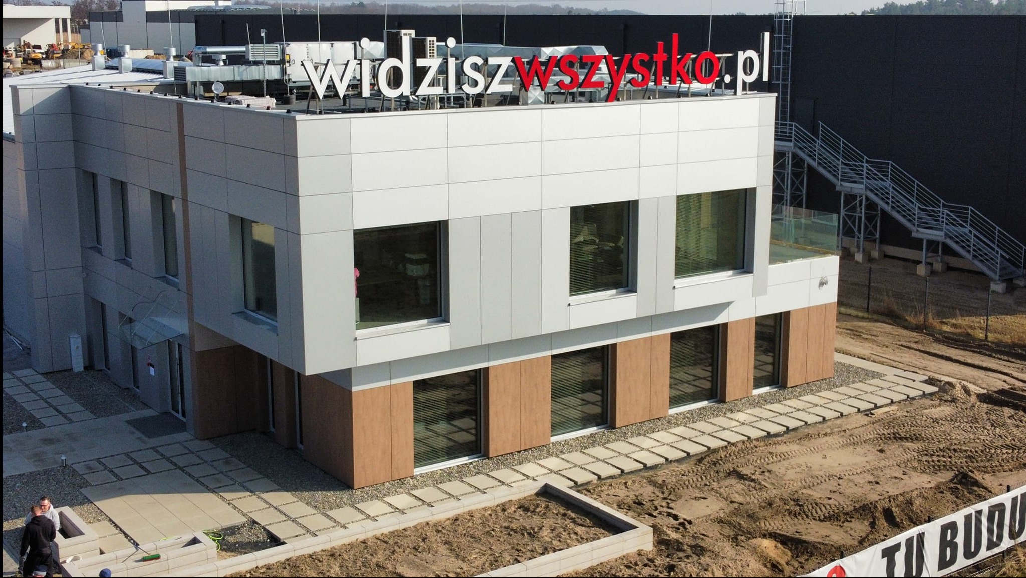 Nowoczesny budynek biurowy z elewacją z paneli w odcieniach szarości i brązu, z napisem 'widziszwszystko.pl' na dachu i zewnętrznymi schodami przeciwpożarowymi.