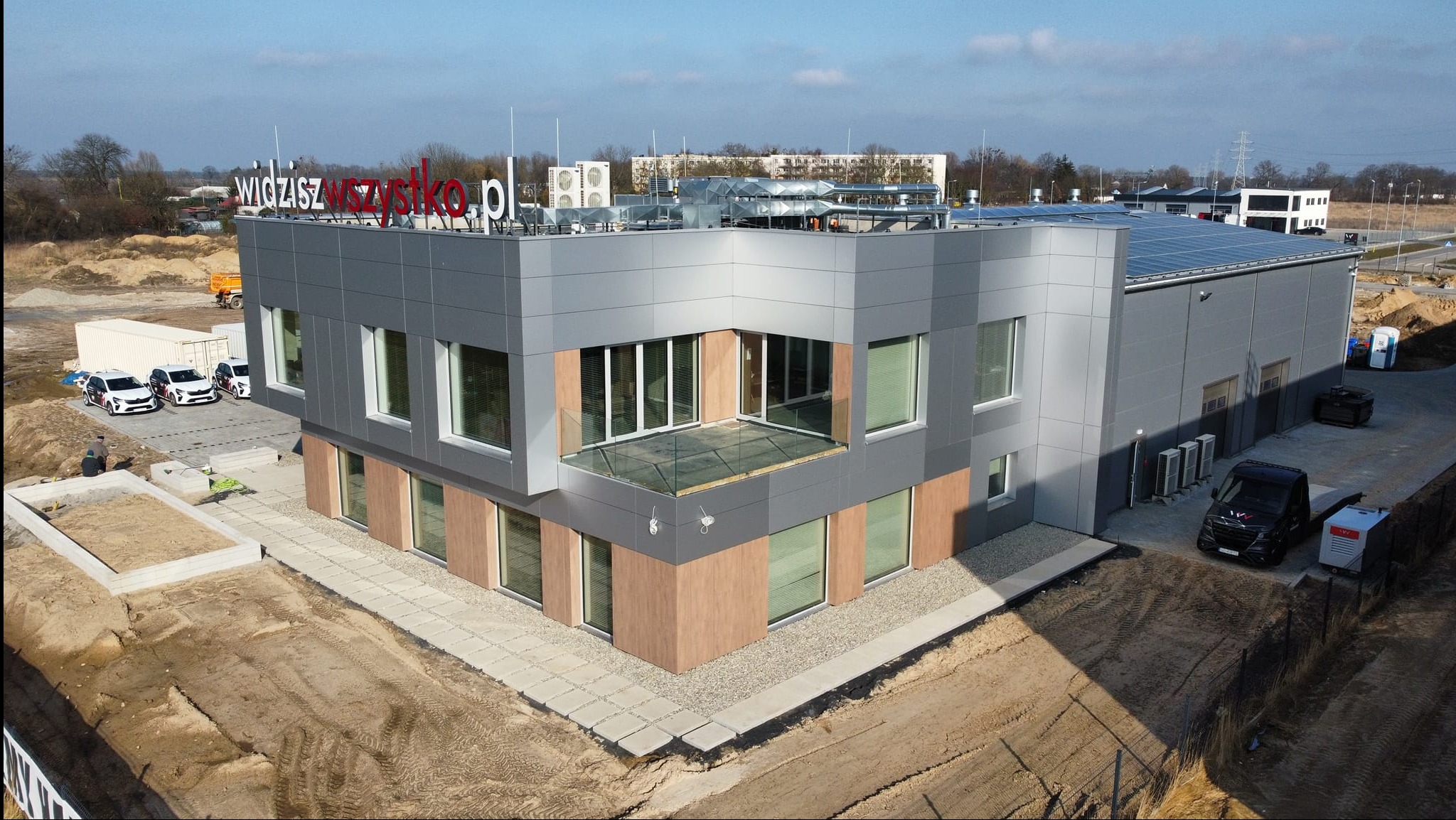 Nowoczesny budynek biurowy z szarą elewacją, drewnianymi akcentami i przeszklonym balkonem, widok z góry na plac budowy i zaparkowane pojazdy.