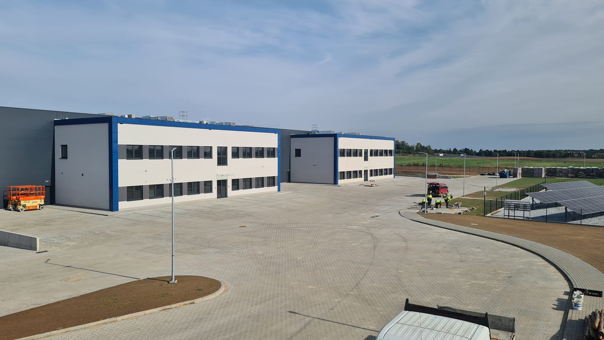 Dwa budynki biurowe z białymi elewacjami i niebieskimi akcentami, widoczne z góry, z placem wyłożonym kostką brukową i ekipą robotników pracujących na zewnątrz, obok instalacji paneli słonecznych.