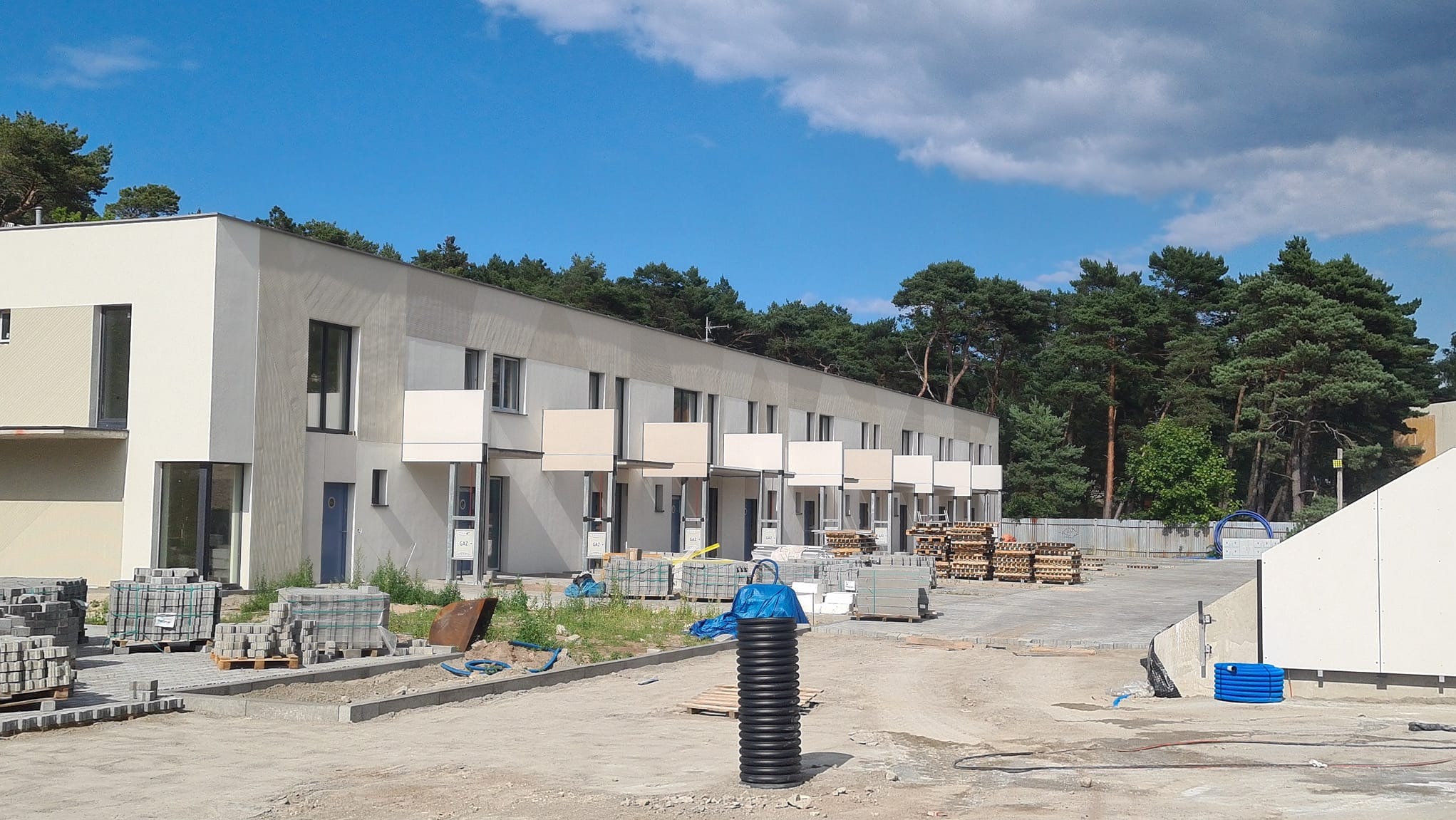 Nowoczesny budynek wielorodzinny w trakcie budowy, widoczne ocieplone elewacje, balkony, materiały budowlane i elementy infrastruktury podziemnej.