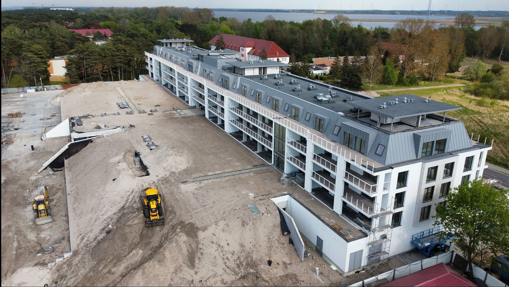 Wielopiętrowy budynek z białymi elewacjami i szarym dachem w trakcie budowy, widoczne dwie koparki na placu budowy, w tle zieleń i zbiornik wodny.