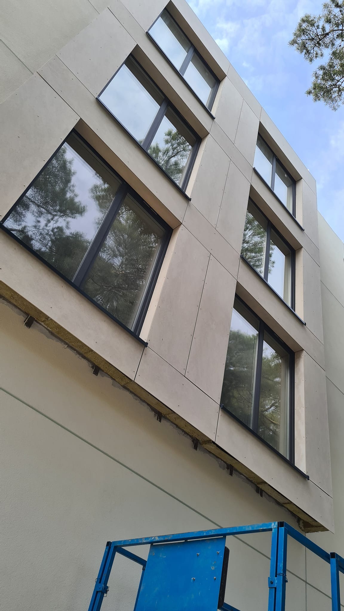 Budynek z elewacją z płyt, widoczne okna z ciemnymi ramami i fragment podnośnika koszowego w dolnej części kadru.