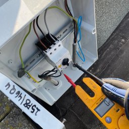 Paweł Michałowski - Elektryk mierzy napięcie na styczniku w skrzynce elektrycznej za pomocą multimetru Fluke, widoczne przewody i szyna DIN.