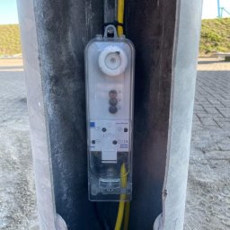 Paweł Michałowski - Zewnętrzna skrzynka elektryczna z bezpiecznikami i okablowaniem, zamontowana wewnątrz metalowego słupka o szorstkiej powierzchni.
