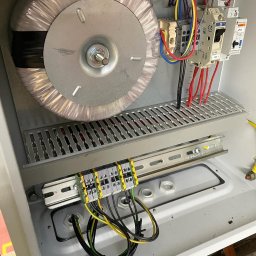 Paweł Michałowski - Wnętrze szafy elektrycznej z transformatorem toroidalnym, bezpiecznikami i listwą zaciskową z podłączonymi przewodami o różnych kolorach.