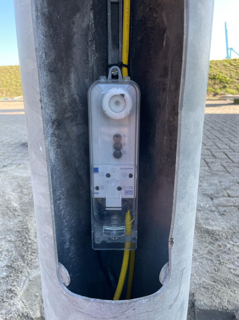 Zewnętrzna skrzynka elektryczna z bezpiecznikami i okablowaniem, zamontowana wewnątrz metalowego słupka o szorstkiej powierzchni.