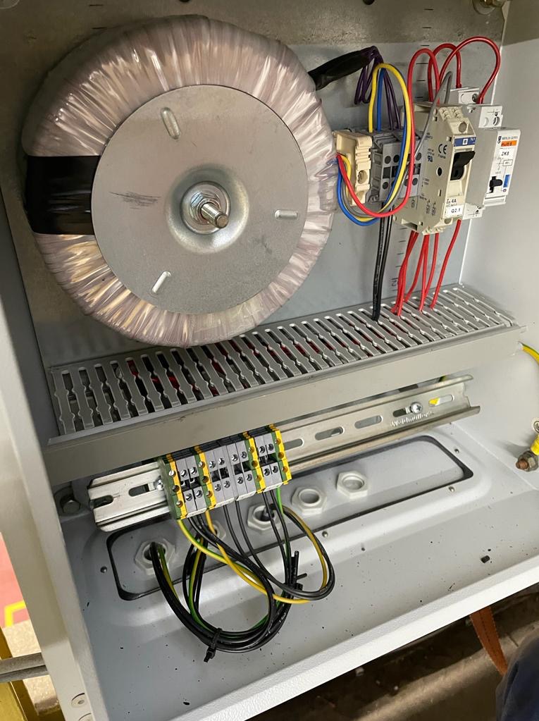 Wnętrze szafy elektrycznej z transformatorem toroidalnym, bezpiecznikami i listwą zaciskową z podłączonymi przewodami o różnych kolorach.