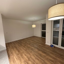 ZG Budownictwo - Puste, jasne wnętrze z drewnianą podłogą, białymi ścianami i dwoma lampami wiszącymi, widok na balkon przez przesuwne drzwi, karton pod drzwiami balkonowymi.