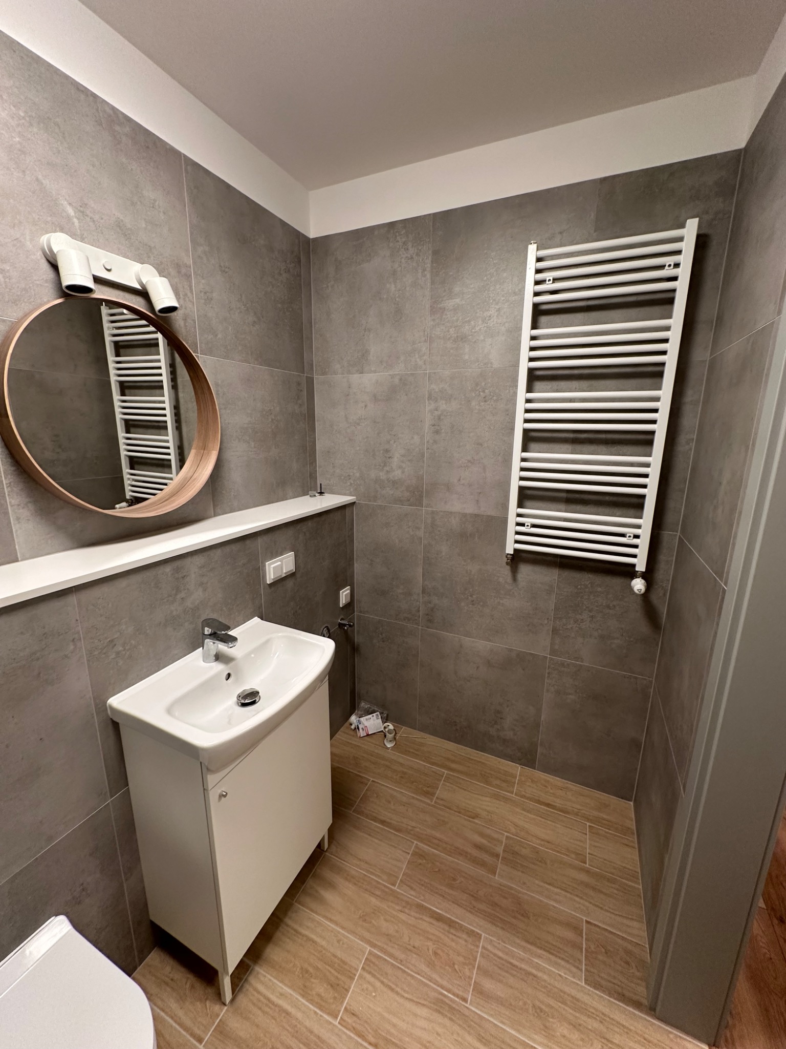 Nowoczesna łazienka z szarymi płytkami, drewnopodobną podłogą, okrągłym lustrem z drewnianą ramą i białym grzejnikiem drabinkowym.