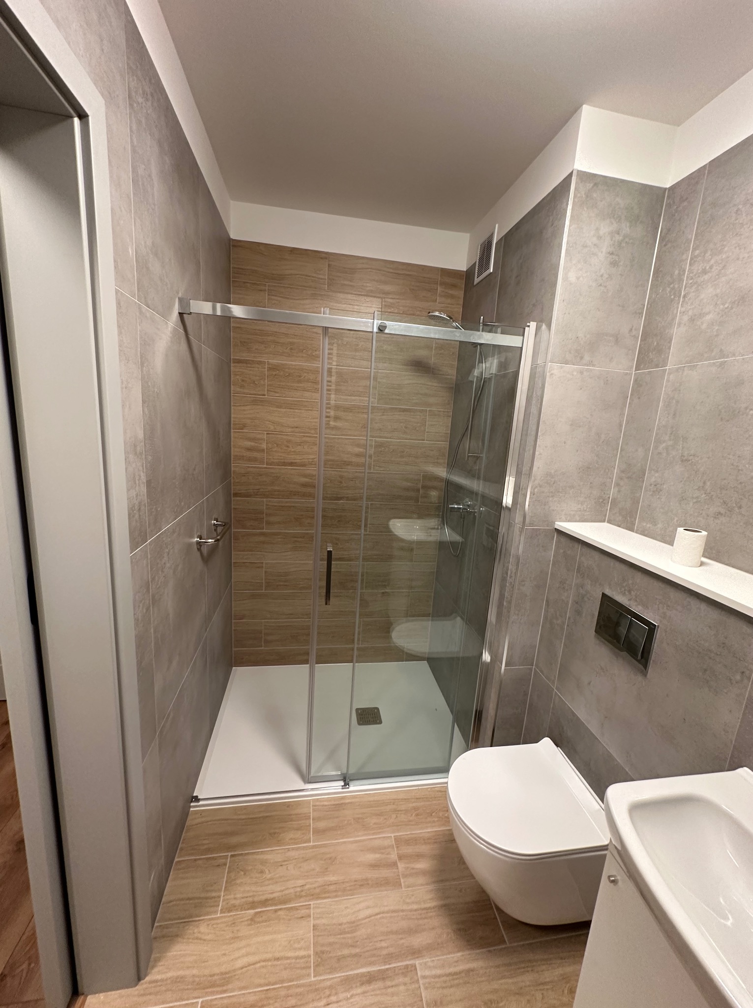 Nowoczesna łazienka z kabiną prysznicową typu walk-in, toaletą podwieszaną i umywalką, wykończona płytkami w odcieniach szarości i drewna, z akcentem w postaci rolki papieru toaletowego na półce.