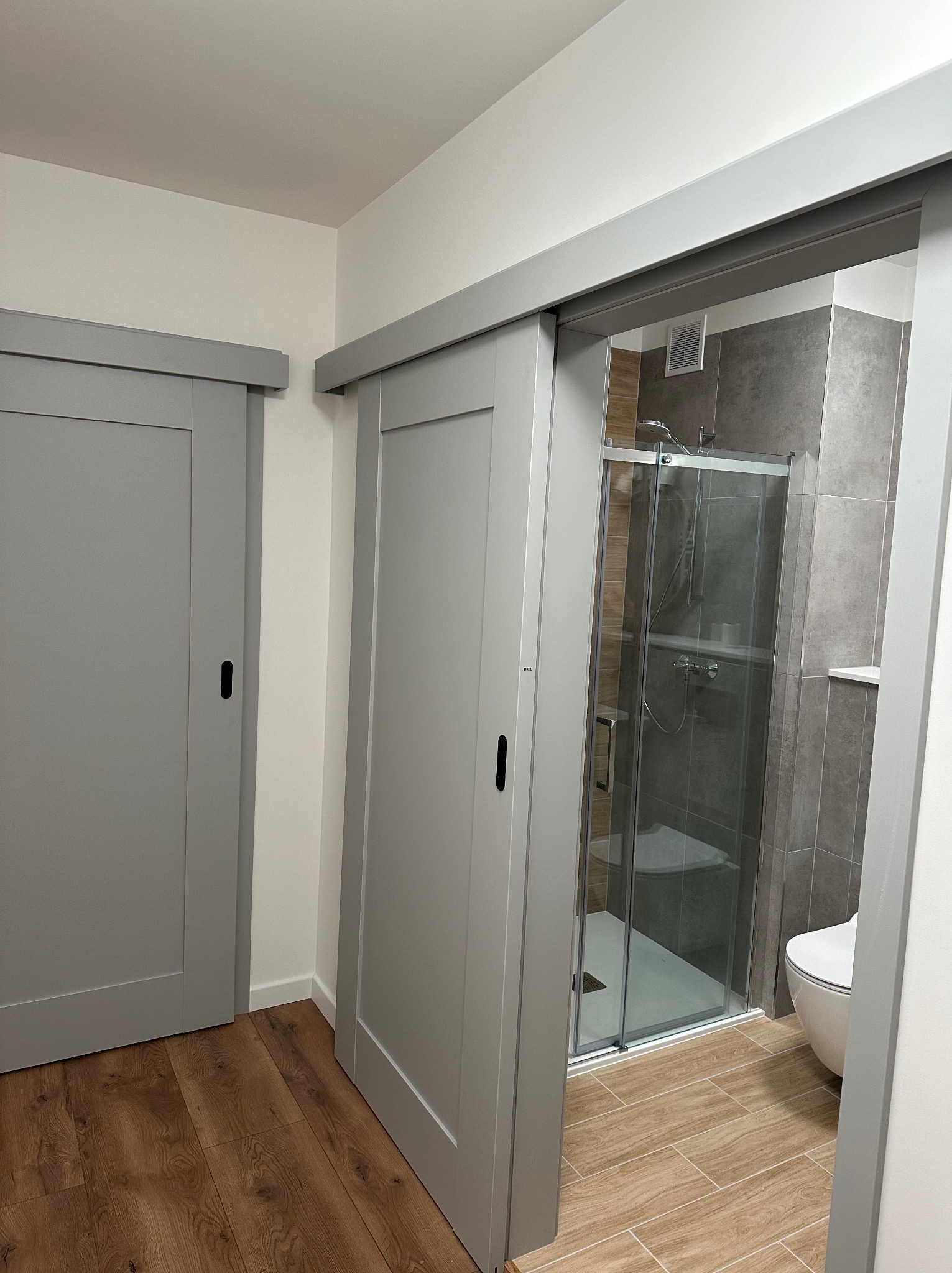 Szare, przesuwne drzwi do łazienki z kabiną prysznicową i toaletą, widoczne panele podłogowe imitujące drewno.