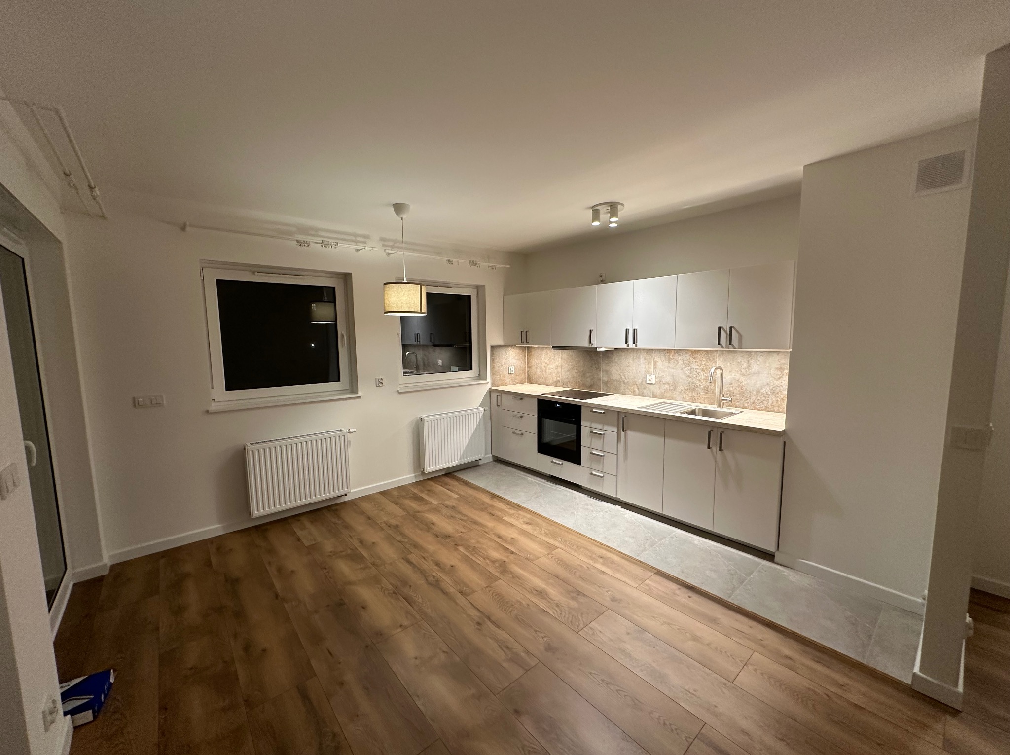 Wykończona kuchnia w nowym mieszkaniu z białymi szafkami, drewnianą podłogą i szarą strefą przy zabudowie kuchennej, widok na okno i grzejniki.