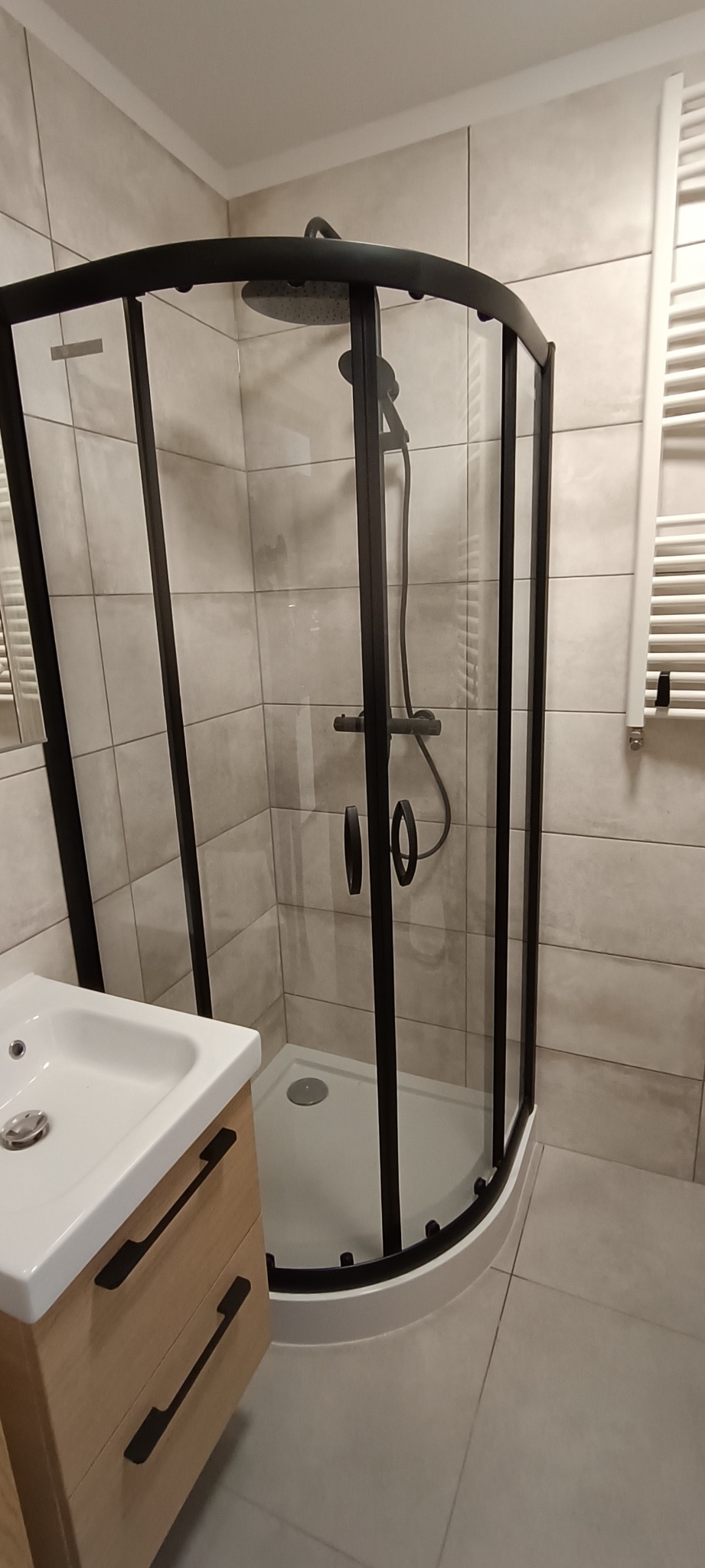 Wyremontowana łazienka z kabiną prysznicową w czarnej ramie, umywalką z szafką z drewnopodobnymi frontami i grzejnikiem drabinkowym na ścianie pokrytej płytkami w odcieniach szarości.