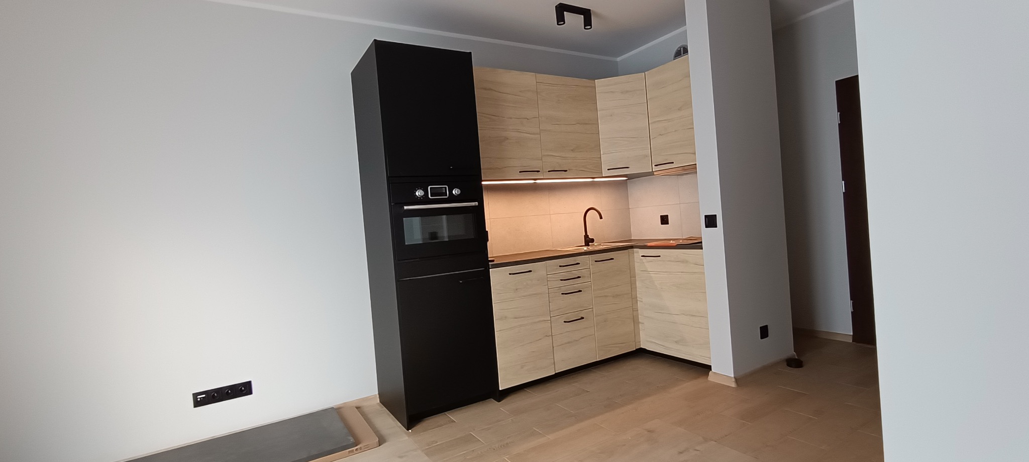 Nowoczesna kuchnia z zabudową w odcieniach beżu i czerni, oświetlenie LED pod szafkami wiszącymi, bateria w kolorze ciemnego brązu, jasna podłoga i szare elementy na podłodze w rogu.