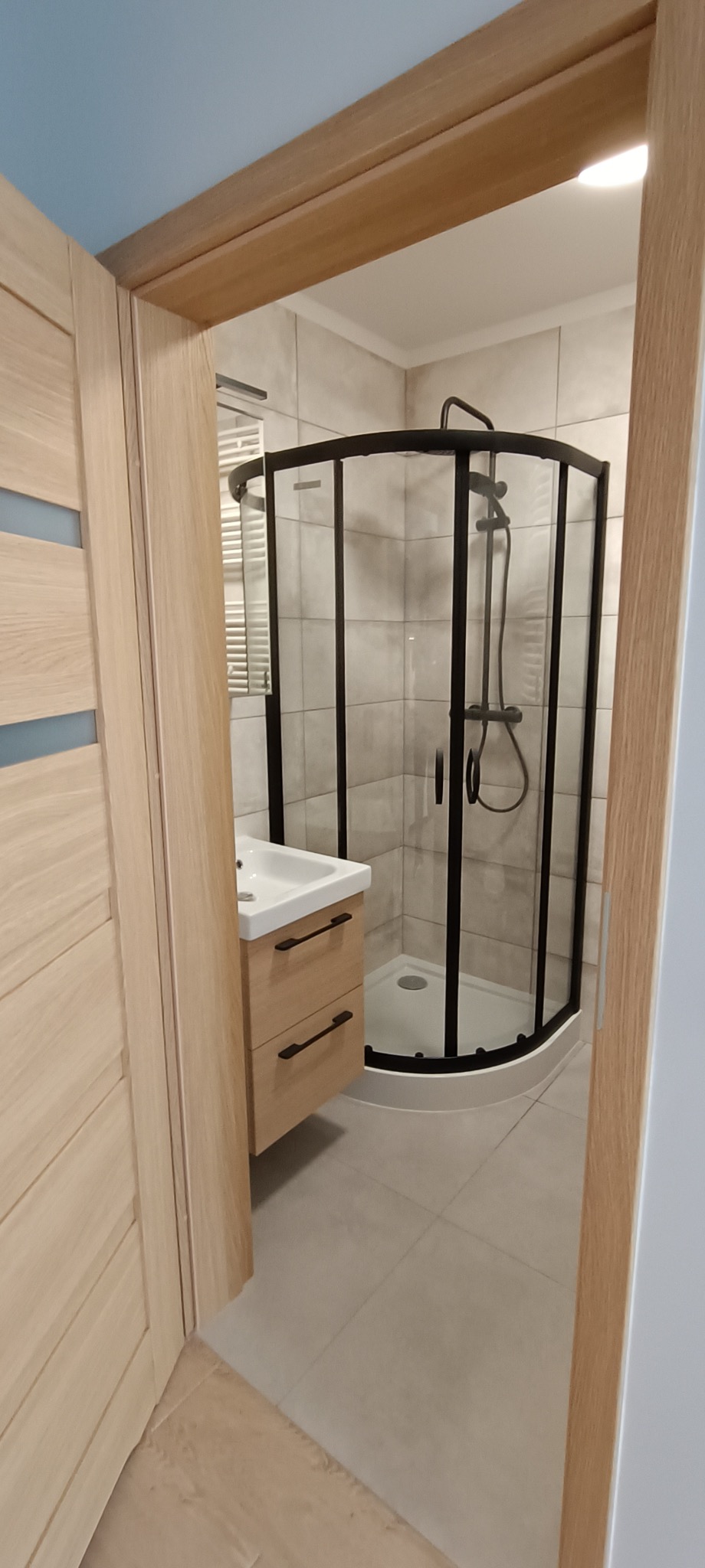 Nowoczesna łazienka z kabiną prysznicową z czarną ramą, umywalką z szafką w odcieniach drewna, wykończona jasnymi płytkami i podłogą. Widok zza drewnianych drzwi z przeszkleniami.