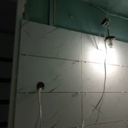 Fragment ściany w trakcie układania białych płytek z szarym wzorem marmuru, widoczne krzyżyki dystansowe, otwór na instalację elektryczną i wisząca żarówka.