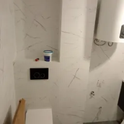 Nowa łazienka z białymi płytkami imitującymi marmur, wiszącą toaletą w kartonie, bojlerem i wnęką z puszką farby oraz pędzlem.