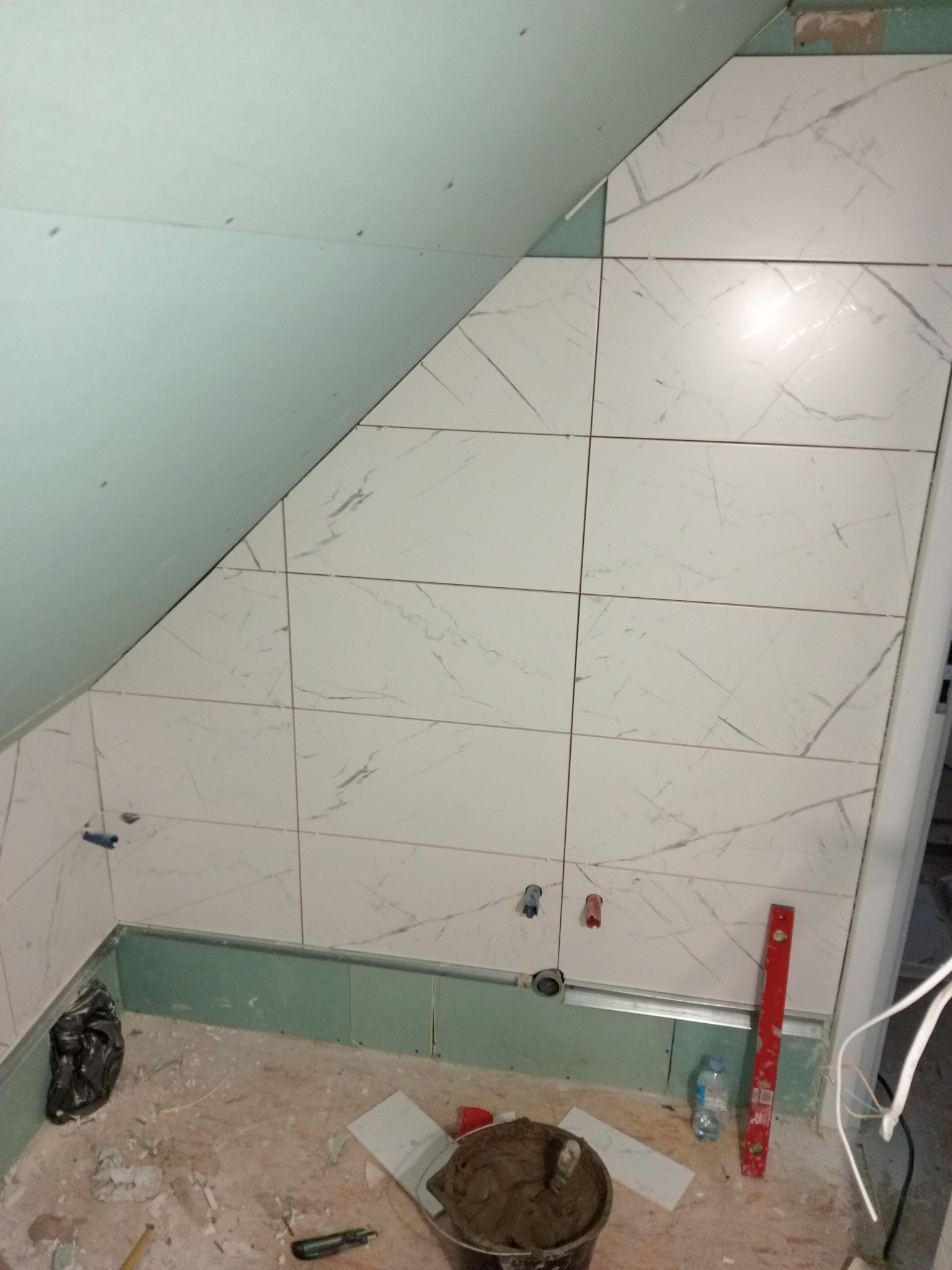 Układanie białych płytek imitujących marmur w łazience na poddaszu, widoczny poziomica, wiadro z zaprawą i fragment surowego wykończenia.