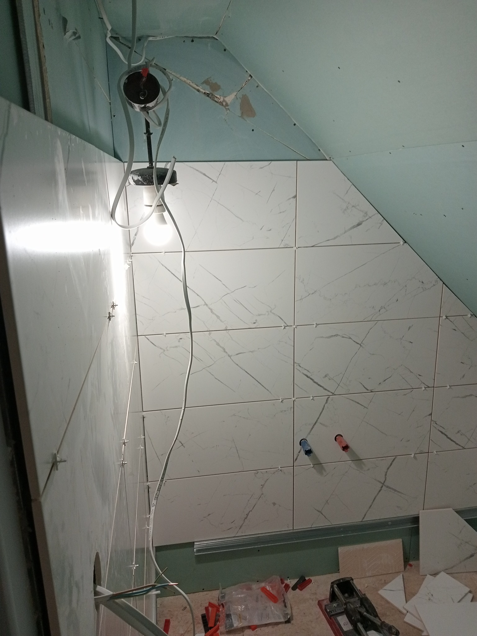 Układanie białych płytek imitujących marmur na ścianie w łazience w Ostródzie, widoczne krzyżyki dystansowe i wystające rury instalacji wodnej, sufit w trakcie wykańczania.