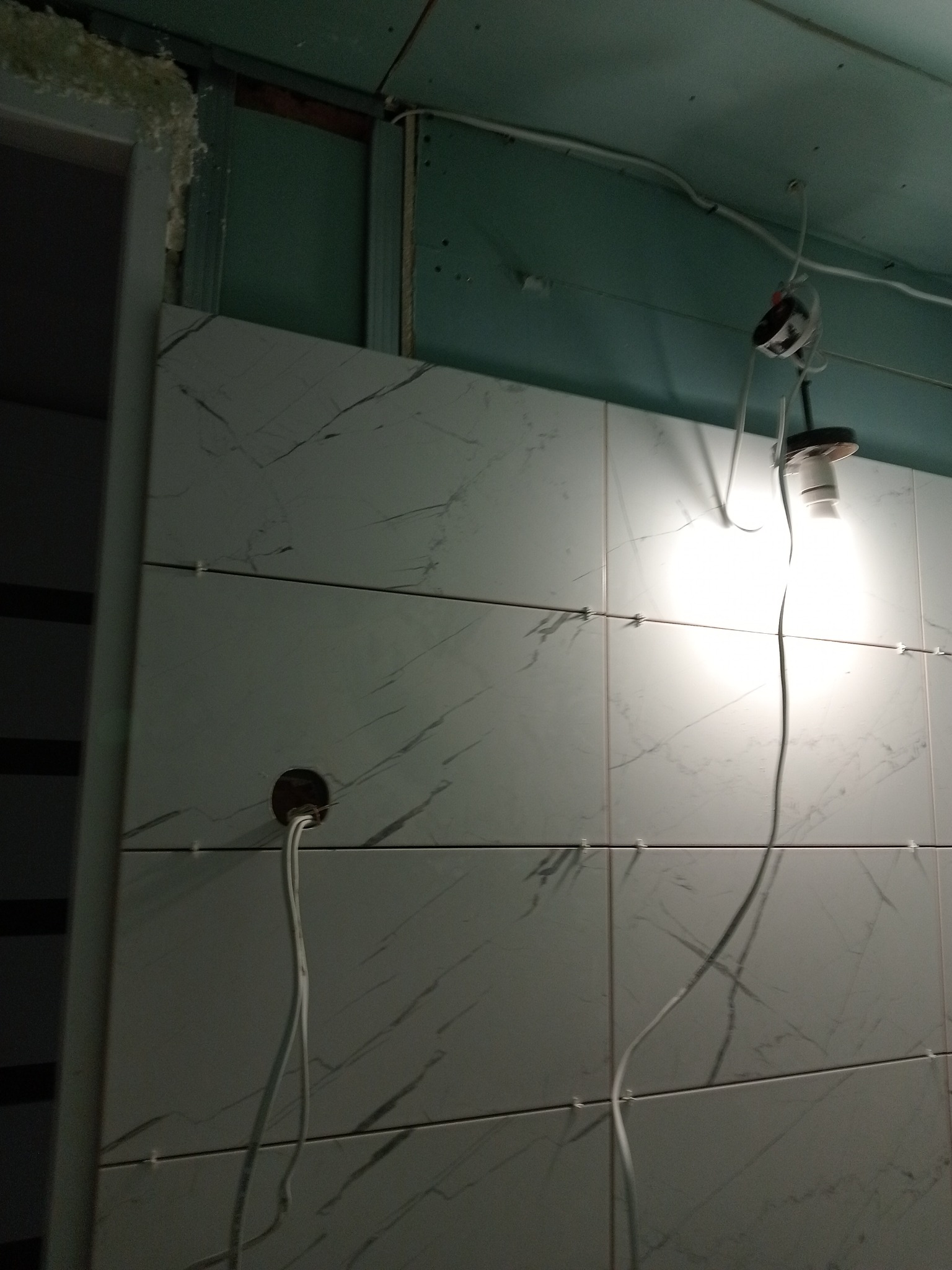 Fragment ściany w trakcie układania białych płytek z szarym wzorem marmuru, widoczne krzyżyki dystansowe, otwór na instalację elektryczną i wisząca żarówka.