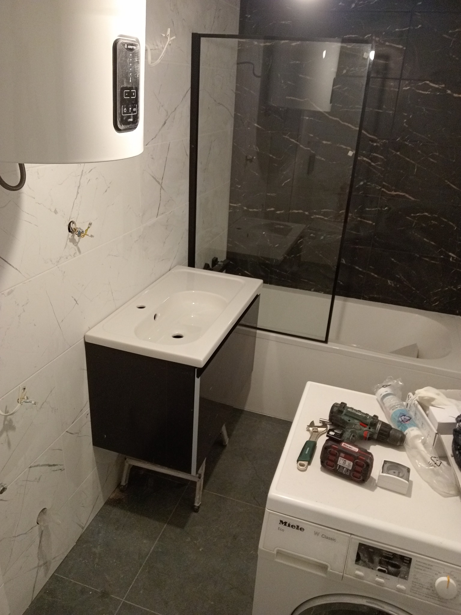 Niedokończona łazienka z białą umywalką na czarnej szafce, białą wanną z czarną szklaną ścianką prysznicową, bojlerem Ariston i pralką Miele, narzędzia na pralce, widoczne niedokończone otwory...