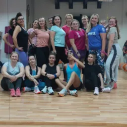 Grupa kobiet w strojach sportowych pozuje do zdjęcia w sali fitness z drewnianą podłogą i widoczną instalacją pod sufitem.