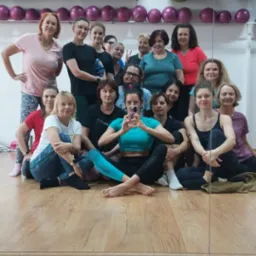 Grupowe zdjęcie z zajęć jogi, kobiety siedzą na podłodze w sali, w tle piłki do ćwiczeń i lustro.