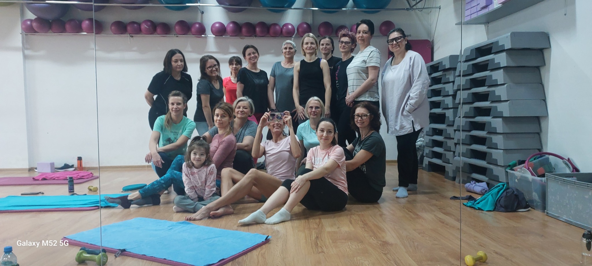 Grupowe zdjęcie uczestniczek zajęć fitness w sali gimnastycznej z lustrem, widoczne maty do ćwiczeń, stepy i hantle.
