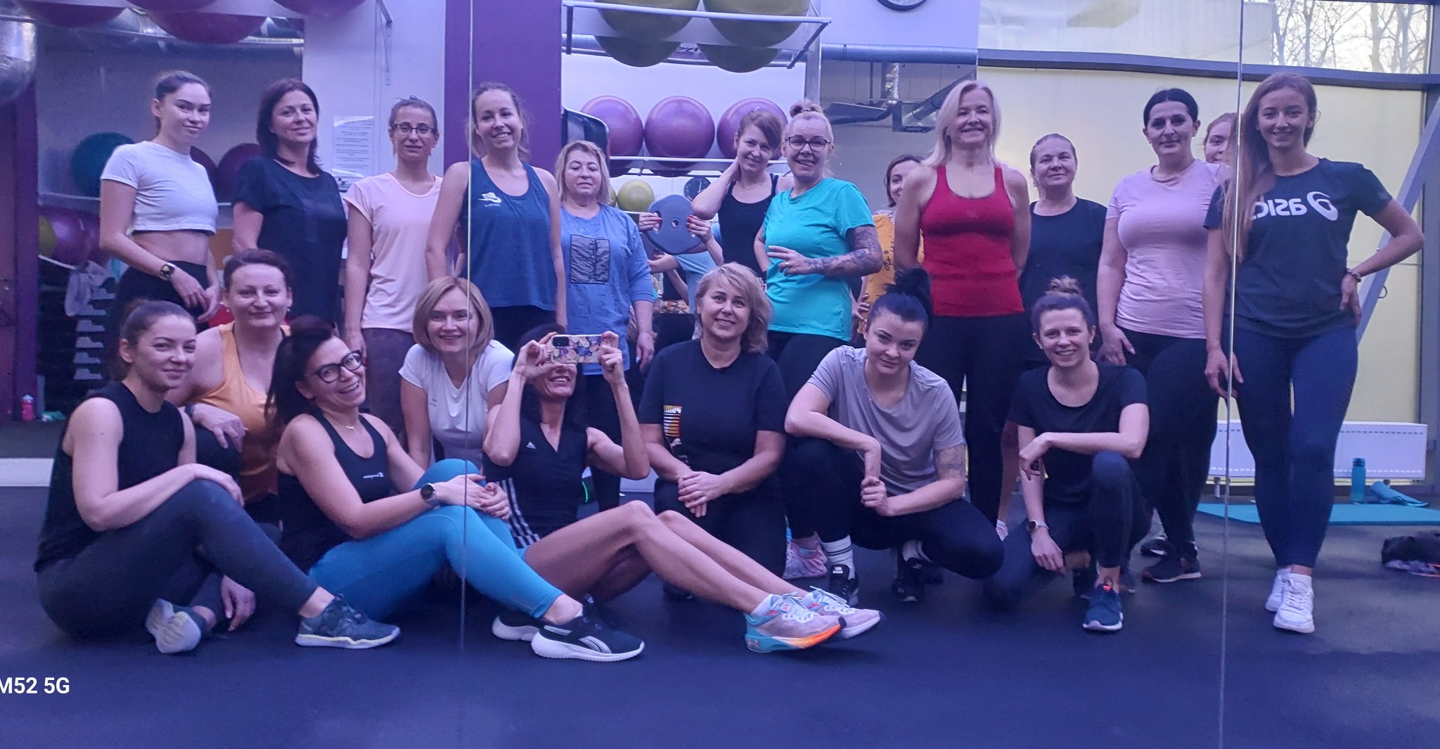 Grupa kobiet w różnym wieku, w strojach sportowych, pozuje do zdjęcia w sali fitness z lustrami i piłkami do ćwiczeń w tle. Jedna z kobiet robi zdjęcie telefonem.
