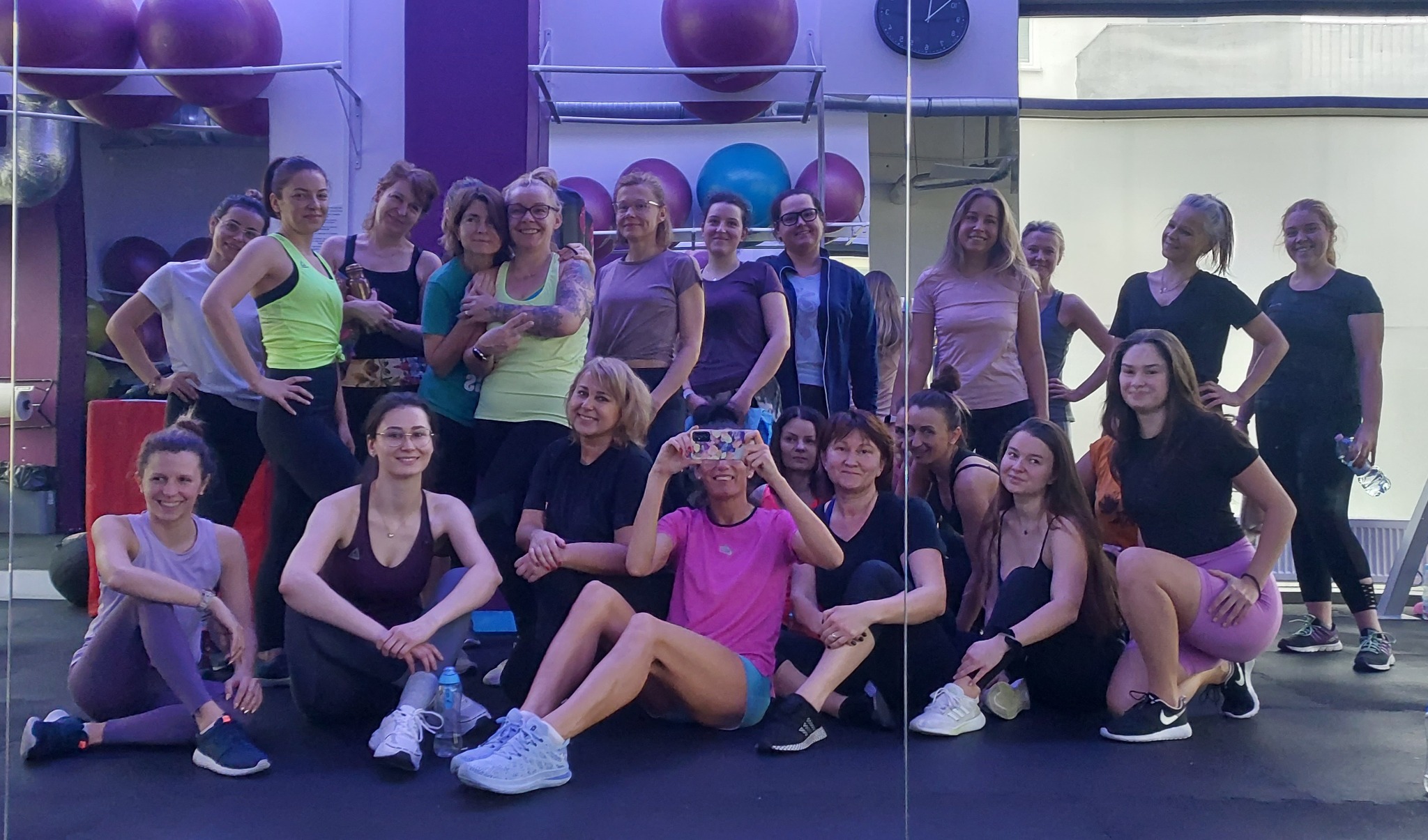 Grupa kobiet w różnym wieku, w strojach sportowych, pozuje do zdjęcia w sali fitness z lustrami i piłkami do ćwiczeń w tle; jedna z kobiet robi im zdjęcie telefonem.