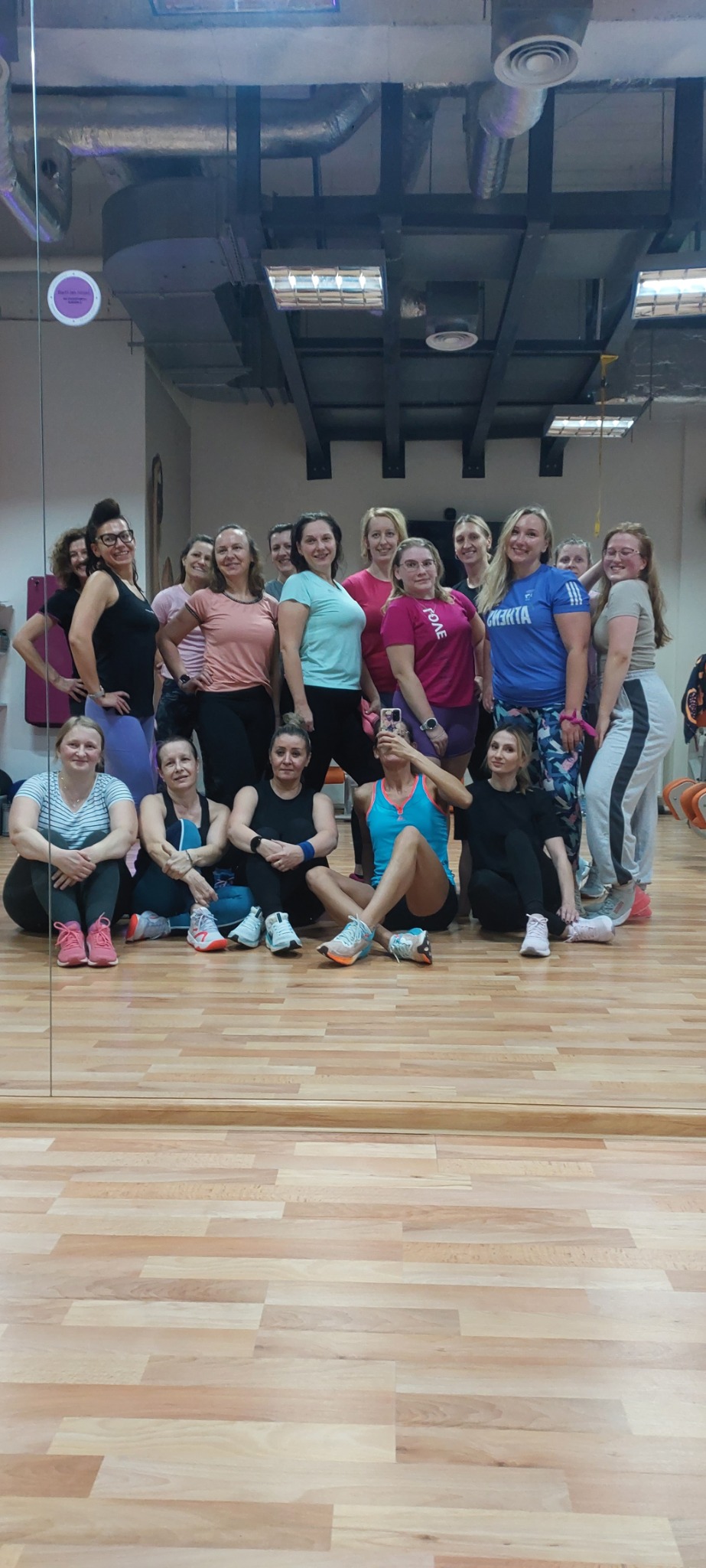 Grupa kobiet w strojach sportowych pozuje do zdjęcia w sali fitness z drewnianą podłogą i widoczną instalacją pod sufitem.
