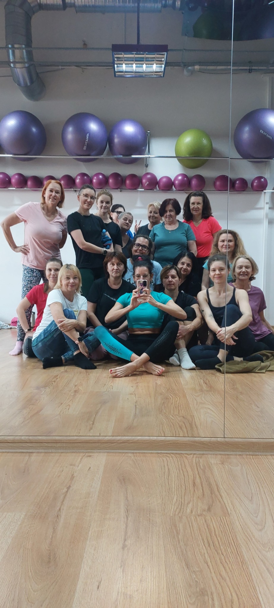 Grupowe zdjęcie z zajęć jogi, kobiety siedzą na podłodze w sali, w tle piłki do ćwiczeń i lustro.