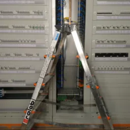 Rozdzielnica elektryczna z licznymi bezpiecznikami i okablowaniem, widoczna drabina aluminiowa z napisem Rolab.