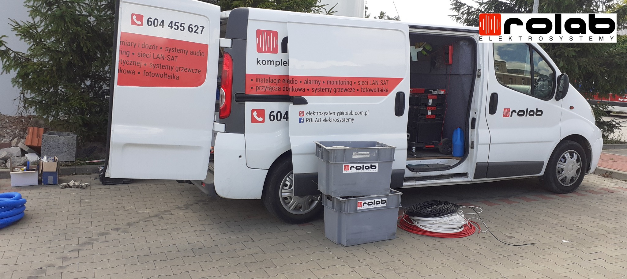 Otwarty furgon firmy elektroinstalacyjnej Rolab z widocznymi narzędziami i przewodami, zaparkowany na chodniku z kostki brukowej, w tle drzewo i gruzowisko.