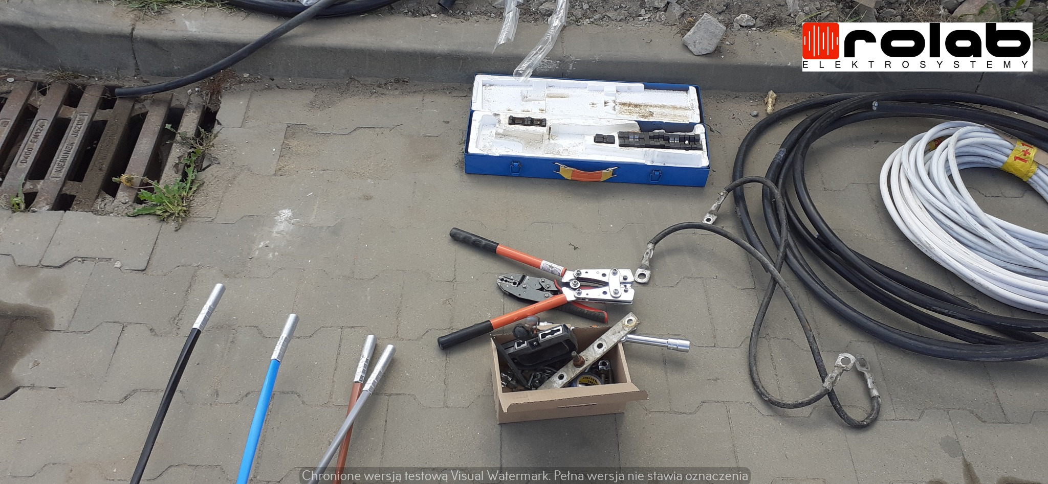 Narzędzia i kable elektryczne rozłożone na chodniku obok studzienki kanalizacyjnej, w tle logo firmy Rolab Elektrosystemy.