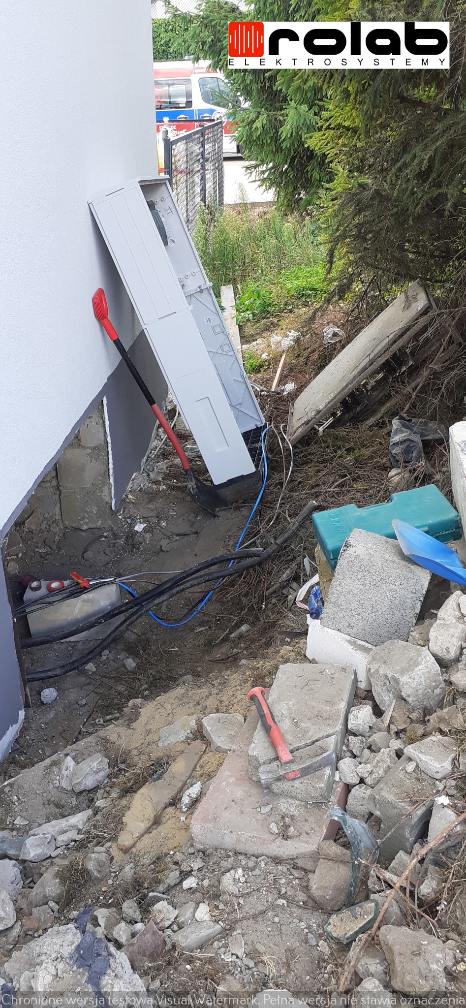 Montaż skrzynki elektrycznej przy ścianie budynku w trakcie prac ziemnych, widoczna łopata, kable i gruz