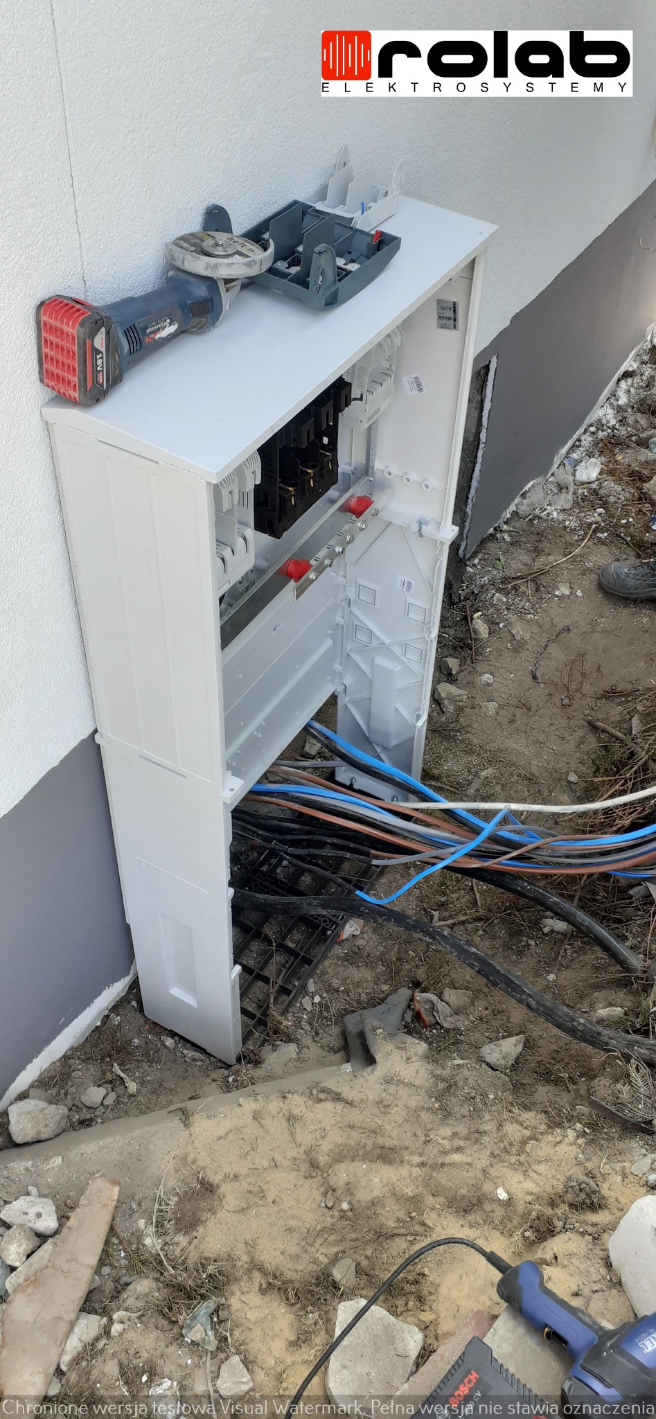 Otwarta, biała skrzynka elektryczna zamontowana na ścianie budynku, z widocznymi przewodami wewnątrz i na zewnątrz, obok leży szlifierka kątowa i wkrętarka akumulatorowa.