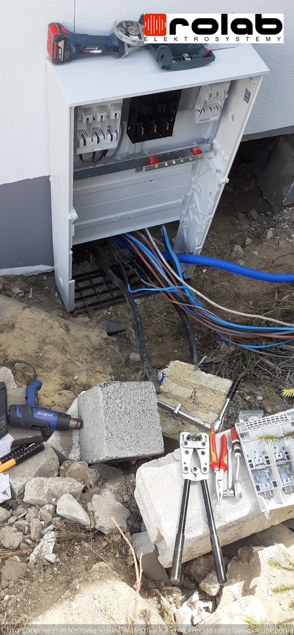 Otwarta skrzynka elektryczna z podłączonymi kablami, narzędzia elektryczne leżące na gruzie i bloczkach betonowych, widoczny szlifierka kątowa na obudowie skrzynki.