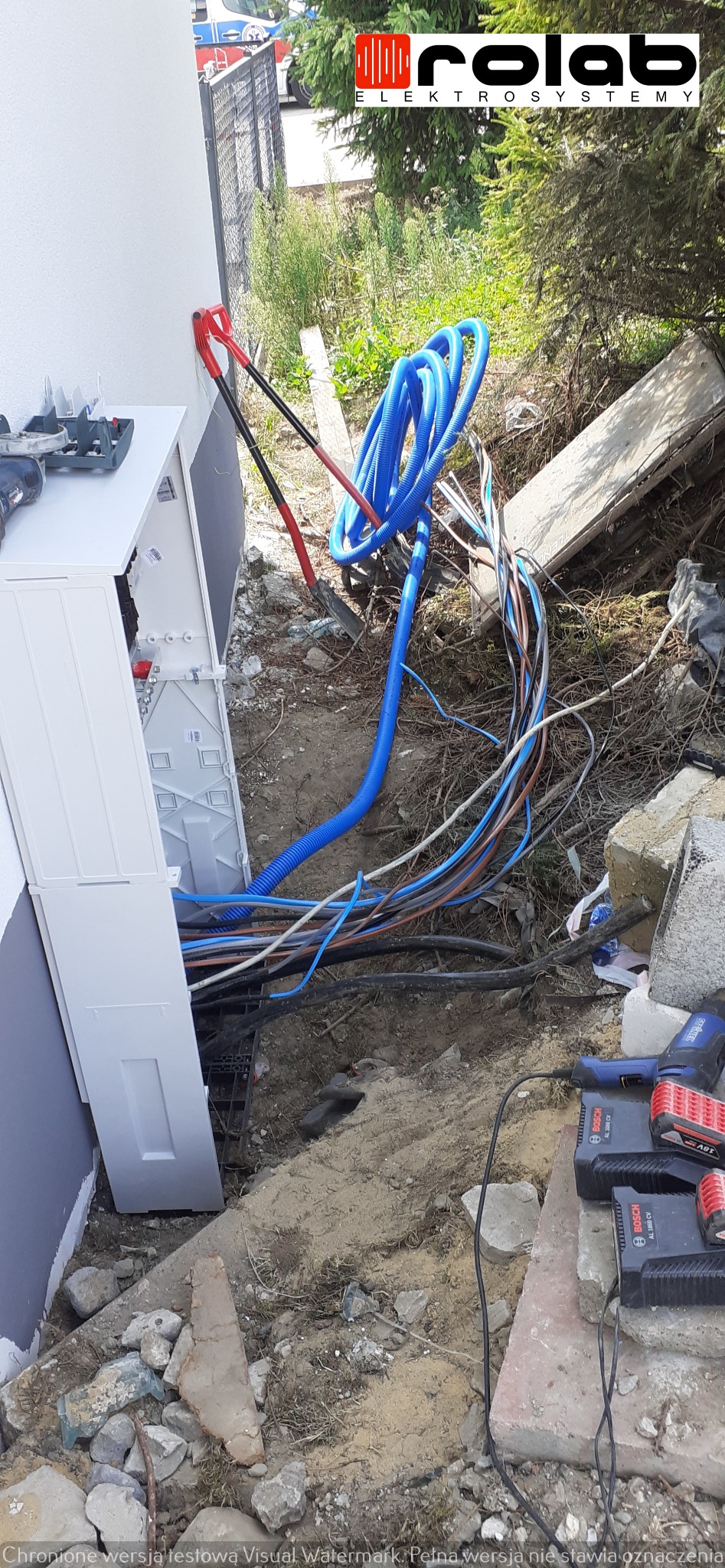 Rozdzielnica elektryczna na zewnątrz budynku w trakcie podłączania, widoczne przewody w peszlach, szpadle i narzędzia akumulatorowe Bosch
