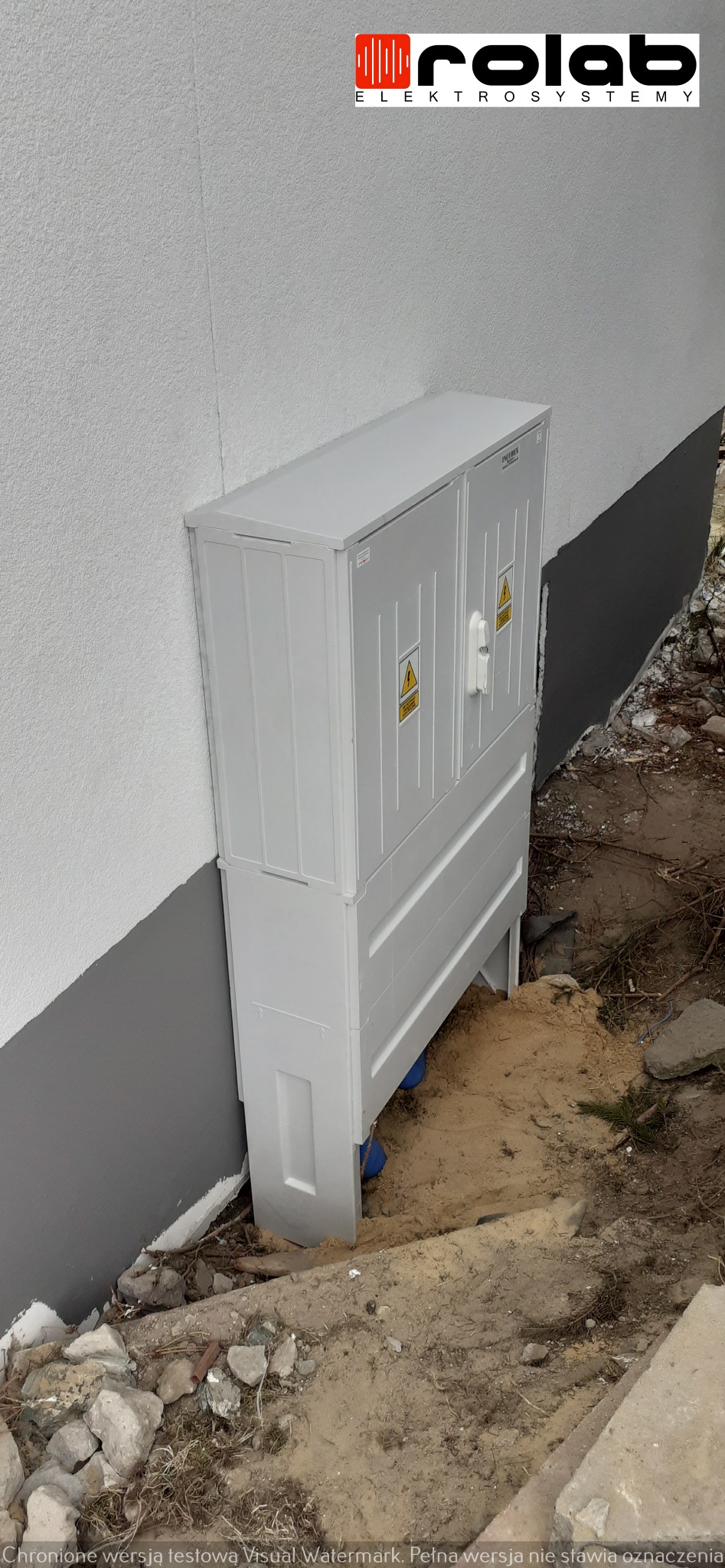 Biała skrzynka elektryczna z symbolem ostrzegawczym wysokiego napięcia, zamontowana na zewnątrz budynku z nierównym terenem.